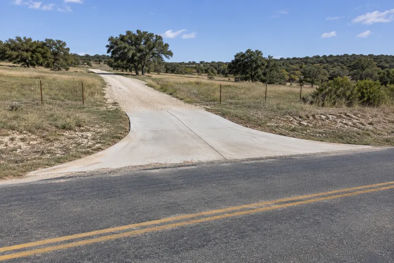A long driveway meeting a two-lane country road through a concrete approach, with open land behind